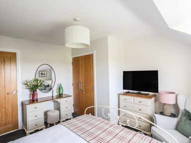 A bedroom with a bed a dresser with a television a desk with a mirror and flowers and a cushioned chair at Dolwaenydd Brynsiencyn