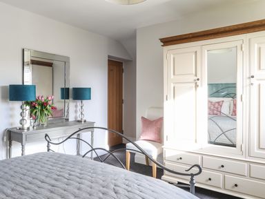 A bedroom with a bed, wardrobe with mirror, dressing table with lamps and flowers, and a chair with a cushion at Dolwaenydd in Brynsiencyn