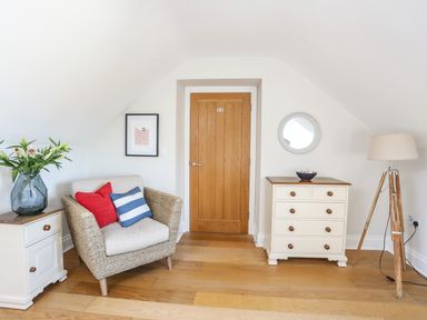 A room with a wooden door a wicker armchair with cushions a white chest of drawers a floor lamp and a side table with a vase Dolwaenydd Brynsiencyn