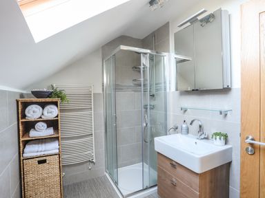 A bathroom with a glass shower enclosure a wooden vanity unit with a white sink rolled towels on a wicker shelf and a skylight at Dolwaenydd in Brynsiencyn