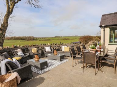 An outdoor patio with wicker chairs and sofas around low tables and a dining table with chairs surrounded by countryside at Dolwaenydd in Brynsiencyn