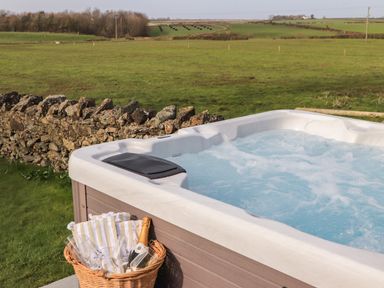 An outdoor hot tub with bubbling water next to a stone wall and a basket with glasses and a bottle in a green field at Dolwaenydd in Brynsiencyn