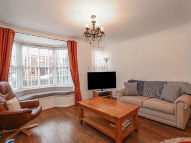 A living room with a beige sofa two cushions a wooden coffee table a leather armchair a bay window with bench seating and red curtains a television on a stand and a chandelier at Job's Cottage in Snettisham