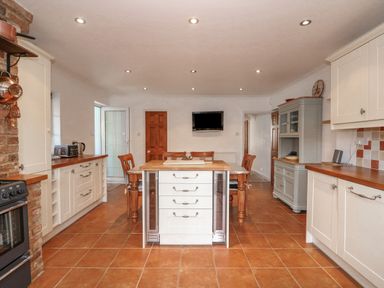 A kitchen with white cabinets a wooden table with chairs a kitchen island a wall mounted tv and tiled floor at Jobs Cottage in Snettisham