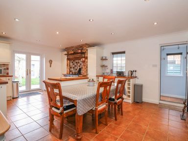 A kitchen with a wooden dining table covered with a polka dot tablecloth and four wooden chairs a brick stove area cabinets and appliances tiled floor and a door leading outside at Job's Cottage in Snettisham