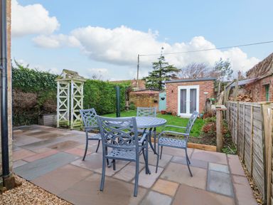 A backyard patio with a metal table and four chairs a small garden shed and greenery at Job's Cottage in Snettisham