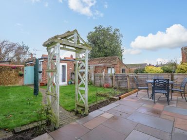 A backyard with a wooden archway a paved patio with a table and four chairs a grass lawn and a brick shed at Job's Cottage in Snettisham