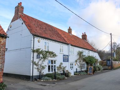 A white building with red tiled roof and multiple chimneys on a street with signs and plants at Jobs Cottage in Snettisham