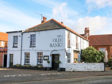 A white two-story building with black windows and a sign reading The Old Bank on a street with plants near the entrance at The Old Barn in Manchester