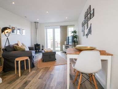 A living room with a gray couch, wooden stool, coffee table, dining table with white chairs, TV, and wood-burning stove at Badgers Way in Tintagel