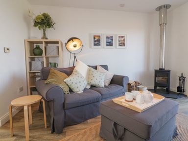 A living room with a grey sofa and cushions a wooden side table a grey ottoman with a tea set a wood burning stove and a shelving unit with decorative items at Badgers Way in Tintagel