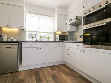 A kitchen with white cabinets stainless steel oven and dishwasher wooden floor and window with blinds at Badgers Way in Tintagel