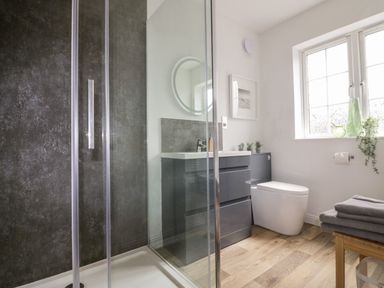 A bathroom with a shower enclosure a gray vanity unit a toilet a window and towels on a wooden stool at Badgers Way in Tintagel