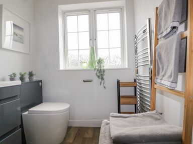 A bathroom with a toilet a window with decorative glass and plants towels on a rack and wooden shelves at Badgers Way in Tintagel