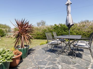 An outdoor patio with a metal table and chairs with cushions and a closed umbrella and potted plants on a stone floor at Badgers Way in Tintagel