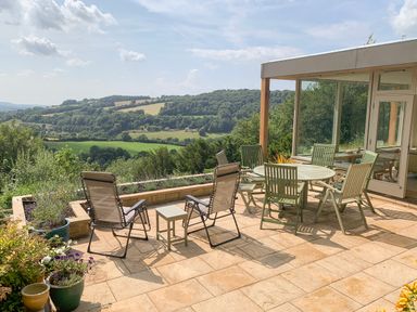 An outdoor patio with green chairs and a table overlooking rolling hills and trees at Under Catswood in Elcombe near Stroud