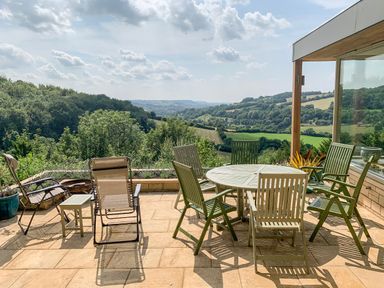A patio with chairs and a round table overlooking a valley at Under Catswood in Elcombe near Stroud