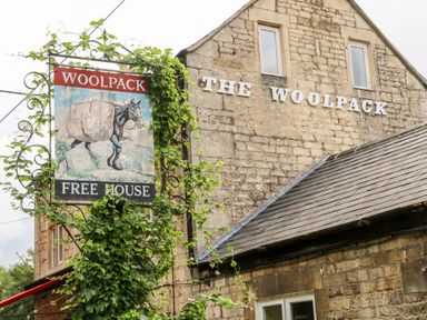 Exterior of a stone building with a sign that reads The Woolpack and a hanging sign with a horse and wool sacks at Under Catswood in Elcombe near Stroud