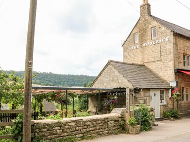 A stone building with the sign The Woolpack a wooden fence and hanging flowers outside at Under Catswood in Elcombe near Stroud