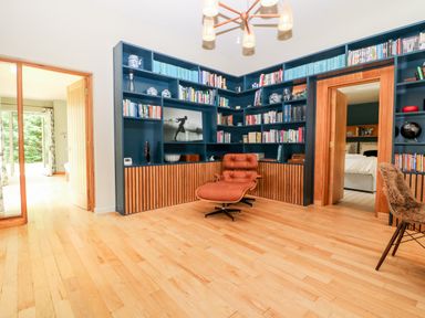 A room with wooden floor built-in blue bookshelves a brown lounge chair and open doors leading to a bedroom and another room at Under Catswood in Elcombe near Stroud