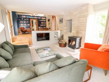 A living room with green sofas an orange armchair a glass coffee table a fireplace and a raised area with a bookshelf at Under Catswood in Elcombe near Stroud