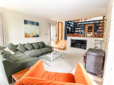 A living room with green and orange chairs a glass coffee table a wall-mounted television and a bookshelf at Under Catswood in Elcombe near Stroud