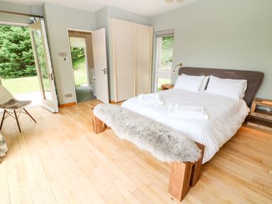 A bedroom with a double bed bedside tables a wooden bench with a fur cover a chair and a door opening to a garden at Under Catswood in Elcombe near Stroud