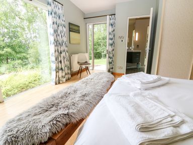 A bedroom with a bed and folded towels a bench with a furry cover a chair and large windows showing a garden at Under Catswood in Elcombe near Stroud