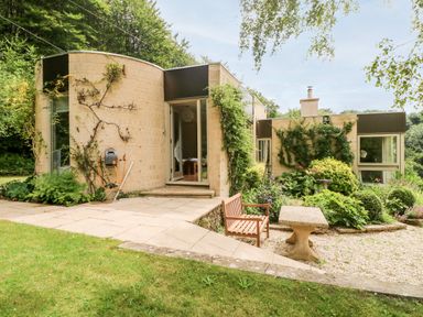 An exterior view of a modern house with a patio garden bench and table surrounded by plants and trees at Under Catswood in Elcombe near Stroud