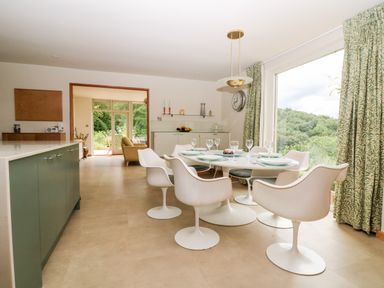 A dining area with a round table set with plates and glasses surrounded by white chairs with large window and green patterned curtains at Under Catswood in Elcombe near Stroud