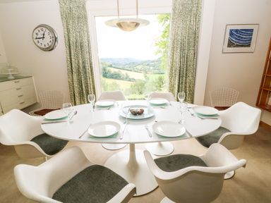 A dining area with a round white table set with plates glasses and cutlery surrounded by white chairs with a large window and patterned curtains in Under Catswood Elcombe near Stroud