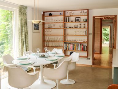 A dining room with a round white table and six white chairs next to a large window and wooden shelves with glassware and books at Under Catswood in Elcombe near Stroud