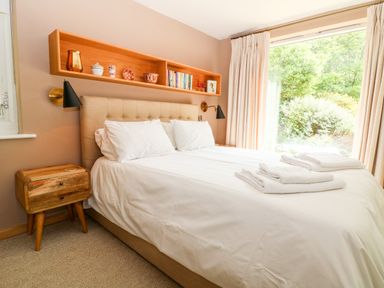 A bedroom with a double bed white bedding wooden bedside table wall shelf with decor and books and a large window at Under Catswood in Elcombe near Stroud