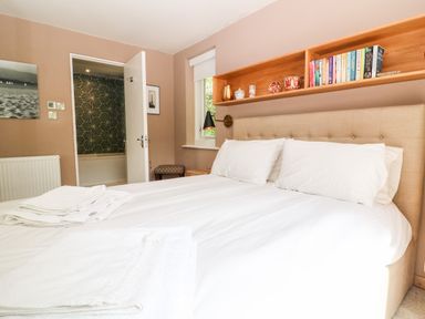 A bedroom with a double bed white linens wooden shelves with books and decor an open door showing a bathroom at Under Catswood in Elcombe near Stroud