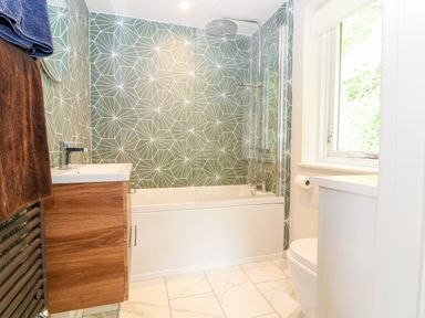 A bathroom with a bathtub and shower, sink with wooden cabinet, towel rack, window, and toilet at Under Catswood in Elcombe near Stroud