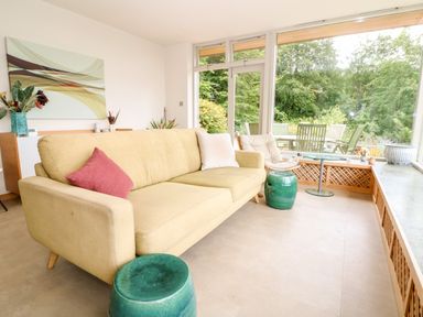 A living room with a beige sofa and green stools by large windows overlooking a garden at Under Catswood in Elcombe near Stroud