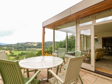 An outdoor patio with a round table and green chairs overlooking a landscape at Under Catswood in Elcombe near Stroud