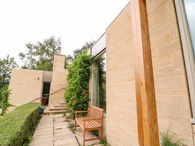 An outdoor patio with a wooden bench against a stone wall and steps leading to a glass door at Under Catswood in Elcombe near Stroud