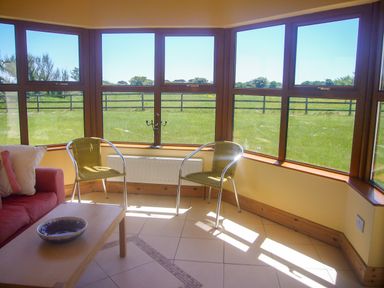 A sunroom with large windows facing a grassy field two chairs a sofa with pillows and a coffee table with a bowl at Tra Gheal in Kilmore County Wexford