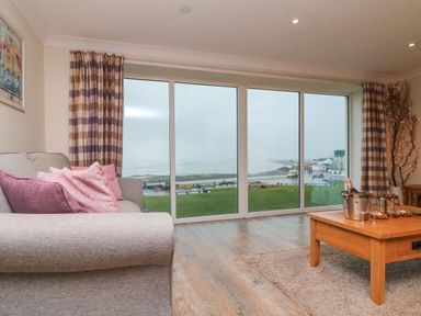 A living room with a gray sofa with pink pillows a wooden coffee table with a champagne bucket and glasses and large windows with a view of a grassy area and coastline at The Lobster Pot in Westward Ho