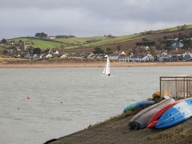 A body of water with a sailboat and kayaks on the shore with houses and green hills in the background at The Lobster Pot in Westward Ho