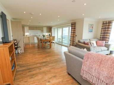 An open plan living room and kitchen with dining table and chairs and sofa with cushions at The Lobster Pot in Westward Ho