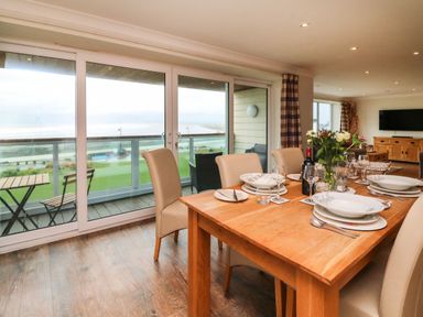 A dining area with a wooden table set with plates glasses and a flower vase next to large glass doors overlooking a balcony with chairs and a view of the sea at The Lobster Pot in Westward Ho