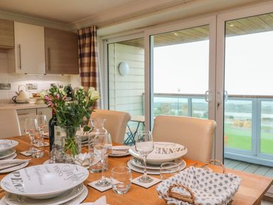 A dining area with a set table including plates wine glasses and a vase of flowers near sliding glass doors overlooking a balcony and outdoor view at The Lobster Pot in Westward Ho