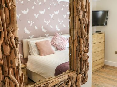 A bedroom with a bed with pillows against a wallpapered wall and a wooden dresser with a television seen through a wooden framed mirror at The Lobster Pot in Westward Ho