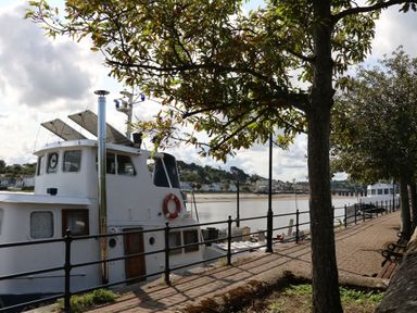 A riverside walkway with trees benches and boats docked by the water at The Lobster Pot in Westward Ho