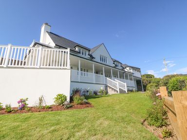 A white house with a large porch and railing overlooking a grass lawn with flower beds at Haul A Gwynt in Criccieth