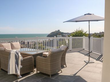 A patio with wicker chairs a glass table and an umbrella overlooking the sea and a distant castle at Haul A Gwynt in Criccieth
