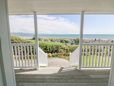 A porch with white railings overlooking a garden with hedges and a view of the sea at Haul A Gwynt in Criccieth