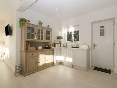 A kitchen with a wooden cabinet filled with glassware next to white cabinets with a toaster kettle and plants at Haul A Gwynt in Criccieth